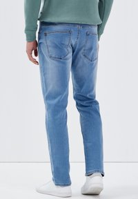 Hellblaue Jeans im Slim-Fit-Stil, mit sichtbaren Gesäßtaschen und Kontrastnähten. Kombiniert mit weißen Sneakers und einem grünen Sweatshirt.