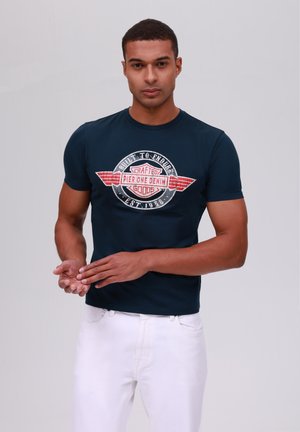 Tricou bleu marin cu guler rotund și un imprimeu grafic cu accente roșii și albe pe piept. Purtați-l cu pantaloni albi.