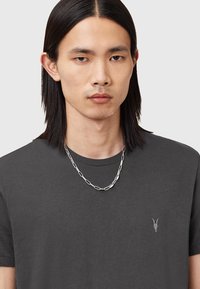 T-shirt grigio a maniche corte con scollatura rotonda e un piccolo design ricamato di teschio di capra a sinistra sul petto. Collana in argento indossata.