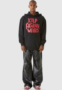 Felpa nera con la scritta "KEEP FASHION WEIRD" in lettere rosa, abbinata a pantaloni larghi neri lucidi e sneakers viola.