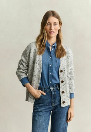 Femme aux longs cheveux blonds porte une chemise en jean bleue, un jean bleu et un cardigan gris tacheté avec de gros boutons, debout devant un fond uni.