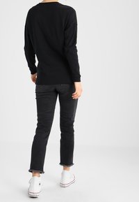 Person steht mit schwarzem Langarmshirt, schwarzen zerrissenen Jeans mit fransigem Saum und weißen High-Top-Converse-Sneakern, mit dem Rücken zur Kamera vor weißem Hintergrund.