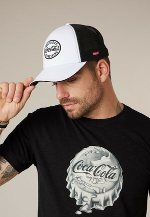 Homme ajustant un bouchon blanc et noir de Coca-Cola, portant un t-shirt noir avec un grand motif argenté de bouchon de bouteille Coca-Cola.