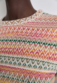 Gros plan sur un pull avec des motifs en zigzag beige, rose, jaune et vert, près du cou et de l'épaule d'une personne.