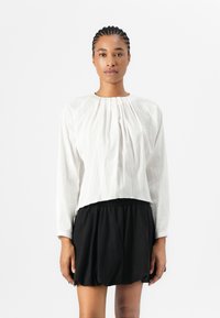 Hvit, teksturert langermet bluse med plissert design. Kombinert med et svart skjørt med elastisk midje. Enkelt halslinje og avslappet passform.