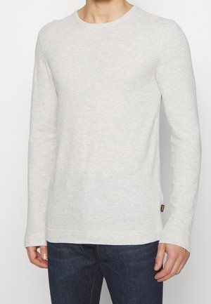 Mann trägt hellgraues langärmliges Rundhalsshirt und dunkelblaue Jeans, steht mit locker an den Seiten hängenden Armen vor einfarbigem Hintergrund.
