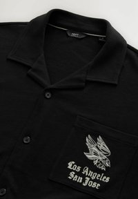 Musta lyhythihainen polo-paita, jossa on kaulus, napit ja brodeerattu design, jossa on kotka ja teksti "Los Angeles San Jose" rinnassa.