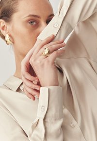 Beige langærmet skjorte med knapmanchetter. Guldøreringe og en ring med et dekorativt design på hånden, der viser glatte teksturer.