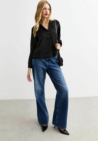 Top lungo a maniche nere in satin con colletto, abbinato a jeans in denim blu a gamba larga e tacchi a punta neri. Borsa a tracolla nera in accompagnamento.