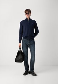 Pulover cu guler tip fular bleumarin, blugi închiși, pantofi negri și o geantă texturată neagră. Modelul poartă ochelari de soare, are un design ajustat și un logo discret.
