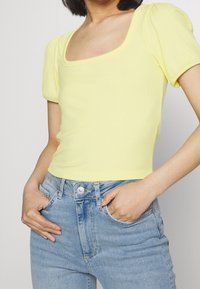 Keltainen ribattu lyhythihainen toppi, jossa on neliön muotoinen pääntie. Tiukka malli, sileä pinta, yhdistettynä vaaleansinisiin farkkuihin, joissa on perinteinen nappisulku.