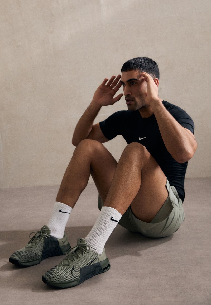 Zapatos deportivos ligeros en verde oliva con una parte superior de malla texturizada, detalles en negro y cordones visibles. Llevados con calcetines blancos y pantalones cortos.