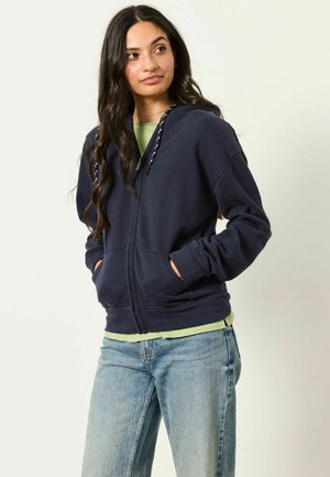 Jeune femme aux longs cheveux foncés portant un sweat à capuche zippé bleu marine, un t-shirt vert clair et un jean bleu, les mains dans les poches, regardant sur le côté.