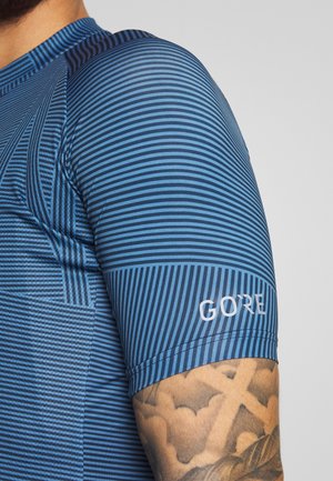 Prawa ręka w geometrycznej niebiesko-paskowanej koszulce z krótkim rękawem, z logo "GORE" nad tatuażem na przedramieniu przedstawiającym chmury i wzór krzyża.