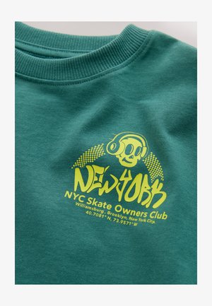 Sinihtävä collegepaita resorikauluksella. Paidassa kirkkaan keltainen kuvitus, jossa on pääkallo kuulosuojaimilla ja teksti "NEW YORK NYC Skate Owners Club."