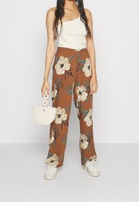 Frau in einem weißen einseitigen Oberteil, braunen weit geschnittenen Hosen mit großem Blumenmuster, weißen Sneakers und einer kleinen weißen Handtasche.