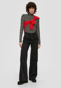 Bluzka z długim rękawem w kwiatowy wzór w kolorze czarnym i beżowym, zestawiona z czarnymi dżinsami z wysokim stanem o szerokich nogawkach oraz czerwonym swetrem przewiązanym w talii.