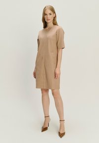 Frau, die in einem beigen knielangen Kleid mit kurzen Ärmeln und braunen spitzen High Heels vor einem schlichten hellen Hintergrund steht.