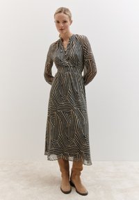 Robe longue et transparente avec un motif géométrique en bleu foncé et beige. Présente un col boutonné et des manches longues. Associée à des bottes marron clair.