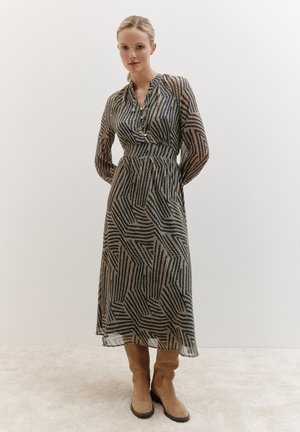 Langes, transparentes Kleid mit einem geometrischen Muster in Dunkelblau und Beige. Verfügt über einen Knopfkragen und lange Ärmel. Kombiniert mit hellbraunen Stiefeln.