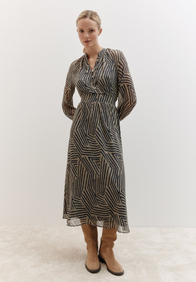 Robe longue et transparente avec un motif géométrique en bleu foncé et beige. Présente un col boutonné et des manches longues. Associée à des bottes marron clair.