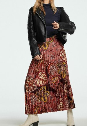 Mujer con chaqueta de cuero negra, suéter de cuello alto azul marino, falda larga roja con estampado floral y botas blancas de tacón, de pie frente a un fondo blanco.