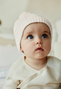 Svetlo roza pletena kapa s pomponom, ki jo nosi malček z modrimi očmi, v kremnem puloverju. Opazne so mehke teksture in preprosti oblikovalski detajli.