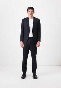 Marineblau gestreifter Anzug mit schlanker Passform, einreihigem Blazer, Reverskragen und flachen Hosen, kombiniert mit einem weißen T-Shirt und schwarzen Schuhen.