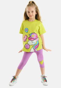 Camiseta verde clara con gráfico de unicornio y acentos coloridos, combinada con leggings morados que tienen un detalle en forma de corazón. Zapatillas plateadas con cordones de arcoíris.