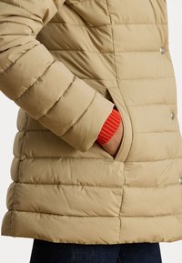Casaco puffer bege com padrão acolchoado horizontal, botões de pressão e bolsos laterais. Punho vermelho vislumbra da manga. Material leve.