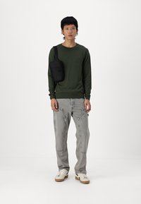 Grønn strikket genser, grå baggy jeans med lommer og beige joggesko. En svart crossover-sekk bæres på skulderen. Enkelt design.