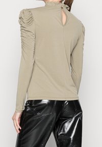 Beige langärmliges Oberteil mit gerüschten Puffärmeln und einem kleinen Schlüsselverschluss mit Knopf auf der Rückseite, kombiniert mit schwarzen glänzenden Lederhosen.