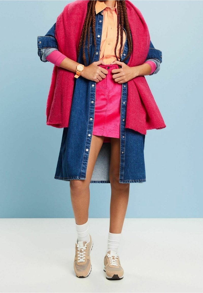Personne portant une jupe rose, une chemise pêche, une longue veste en jean, une écharpe rose, des baskets beige et des chaussettes blanches avec des cheveux tressés visibles.