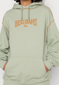 Sudadera verde claro con un bolsillo tipo canguro, que presenta texto bordado en naranja "BDG JEANS" y bordes cosidos en contraste. Ajuste suave y relajado.