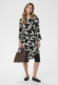 Femme portant une robe florale noire et blanche, des leggings noirs, des ballerines noires, et tenant un grand sac à main marron.