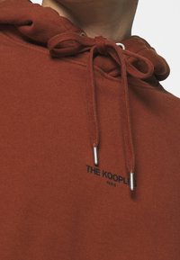 Hoodie i rostfärg gjort av mjukt tyg, med huva som har dragsko med metallspetsar och en svart logotyp tryckt på bröstet.