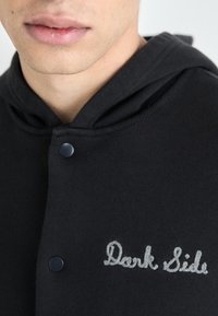 Svart hettegenser med snøring, med trykknapp-feste. "Dark Side" brodert i lys grå på brystet. Glatt, strukturert stoff.