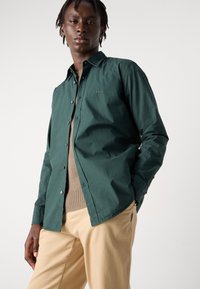 Camisa verde de botonadura con una textura suave y un ligero brillo, que presenta un logo en el pecho y un cuello. Combinada con pantalones beige.