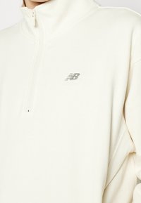 Gros plan d'une personne portant un sweat New Balance écru à fermeture quart de zip, col montant et logo sur la poitrine.