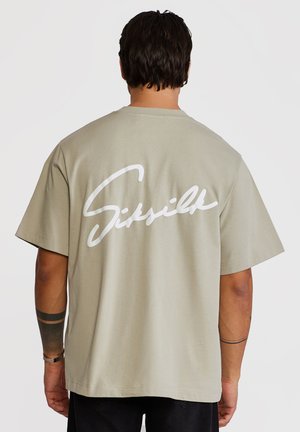 Mand med mørkt hår iført beige oversized t-shirt med hvid kursiv tekst "Sixville" på ryggen, sorte bukser og armtatoveringer.