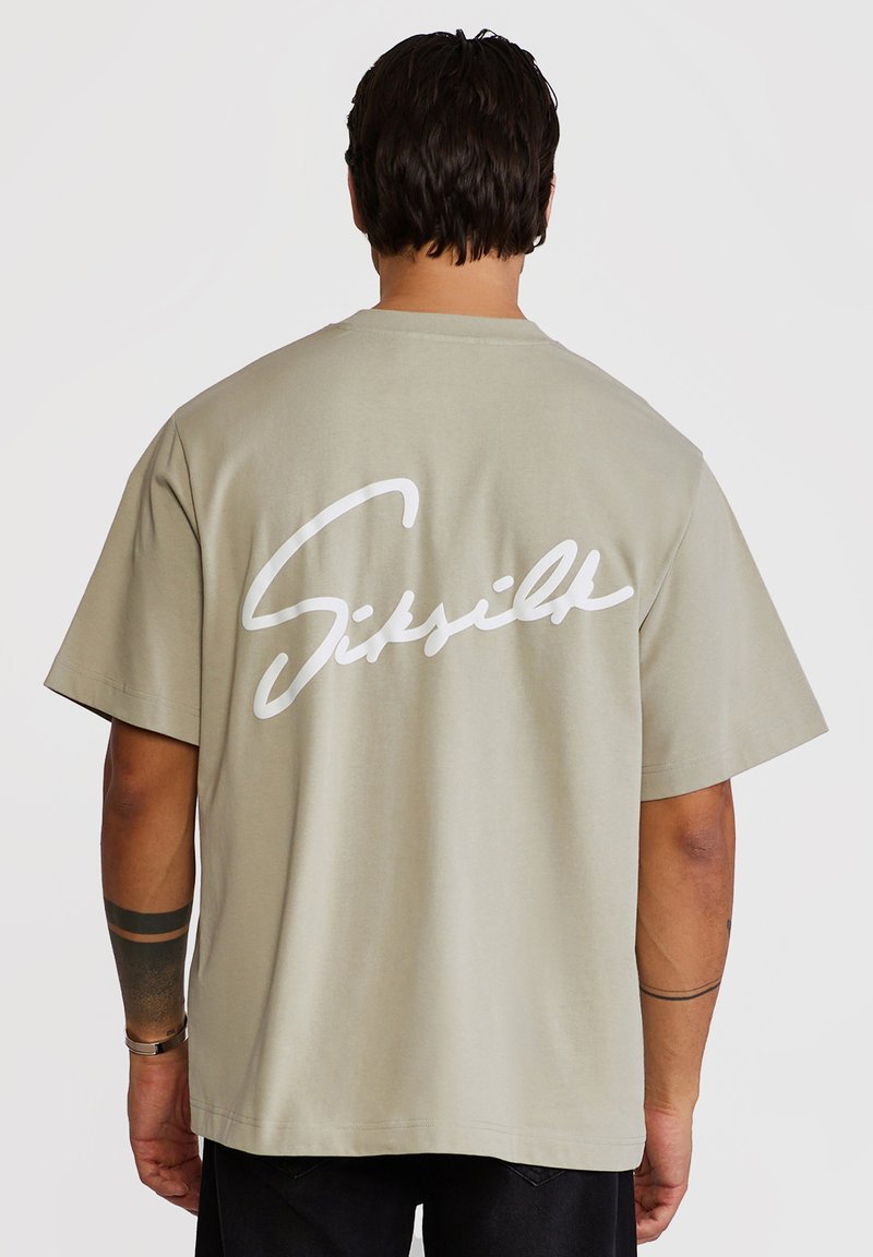 Homme aux cheveux foncés portant un t-shirt beige surdimensionné avec le texte cursif blanc "Sixville" au dos, un pantalon noir et des tatouages sur les bras.