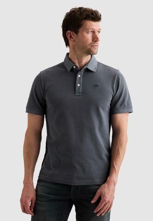 Hombre que lleva una camiseta polo gris oscuro de manga corta y jeans, mirando de lado contra un fondo claro y liso.