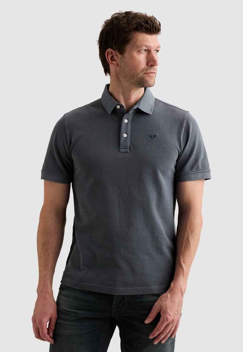 Homme portant un polo manches courtes gris foncé et un jean, regardant sur le côté devant un fond clair uni.