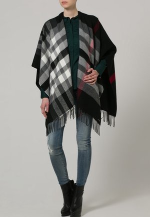 Kobieta ubrana w czarno-biało-szary kraciasty szal z frędzlami na zielonej bluzce, dopasowane dżinsy i czarne botki, stojąca na jednolitym tle.