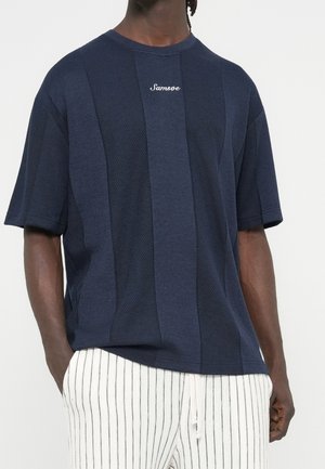 Hombre que lleva una camisa de manga corta texturizada azul marino con el logo "Samsøe" y pantalones blancos con finas rayas verticales negras.
