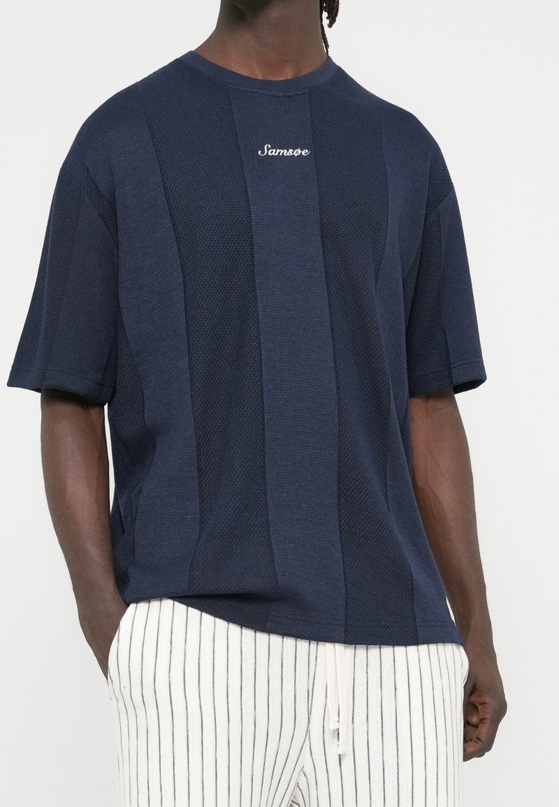Homme portant une chemise bleu marine texturée à manches courtes avec le logo "Samsøe" et un pantalon blanc à fines rayures verticales noires.