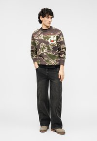 Φούτερ με καμουφλάζ σε αποχρώσεις ελιάς και καφέ, με λευκές και κόκκινες λεπτομέρειες, συνδυασμένο με σκούρο, φαρδύ τζιν και παπούτσια σε απόχρωση ταοπέ.