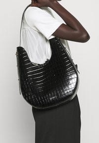 Czarna torebka na ramię z lakierowanej skóry, o lekko zakrzywionym kształcie, z teksturą krokodyla i srebrnymi akcentami.