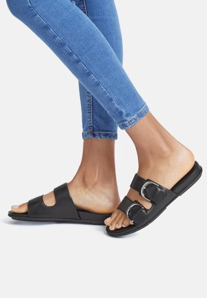 Black slide sandals with two wide straps and silver buckles, featuring an open toe design and flat rubber sole. Jeans partially visible.