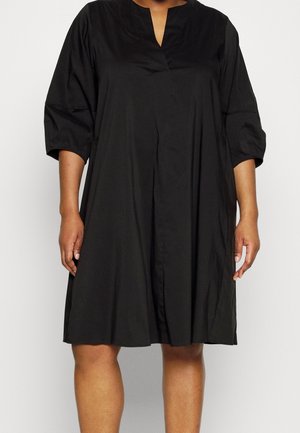 Femme vêtue d'une robe noire ample longueur genou avec des manches trois-quarts et une petite ouverture en V sur un fond clair.